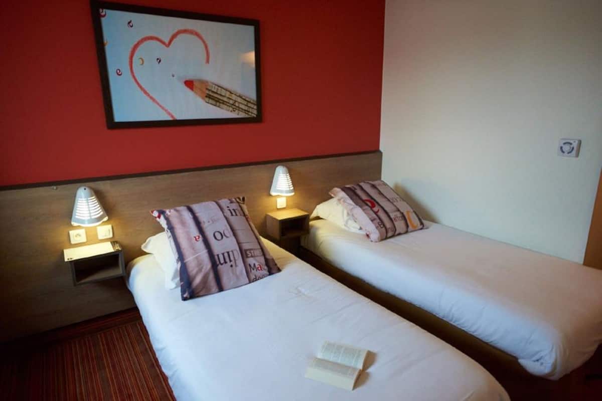 Twee eenpersoonsbedden met nachtkastjes, elk met een lamp, in een kamer met een rode muur en een ingelijst kunstwerk.