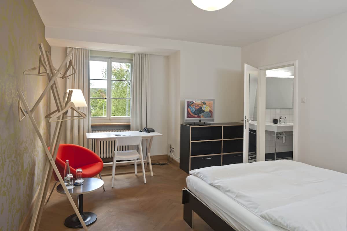Une chambre d’hôtel comprenant un lit, un bureau avec une chaise, une chaise rouge, une télévision, un miroir et une salle de bain avec un lavabo et des toilettes.