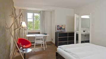 Habitación de hotel con cama, escritorio y silla, una silla roja, televisión, espejo y baño con lavamanos e inodoro.