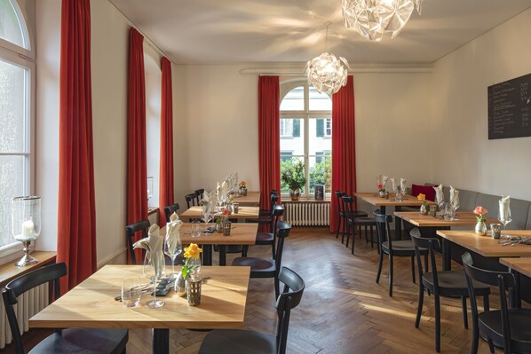 A restaurant with wooden tables and black chairs, red curtains, and a chandelier.