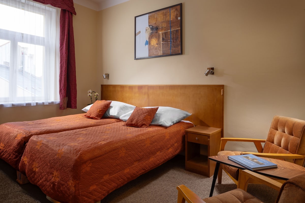 A hotel room with two beds, a wooden headboard, a nightstand, a chair, and a table with a book.