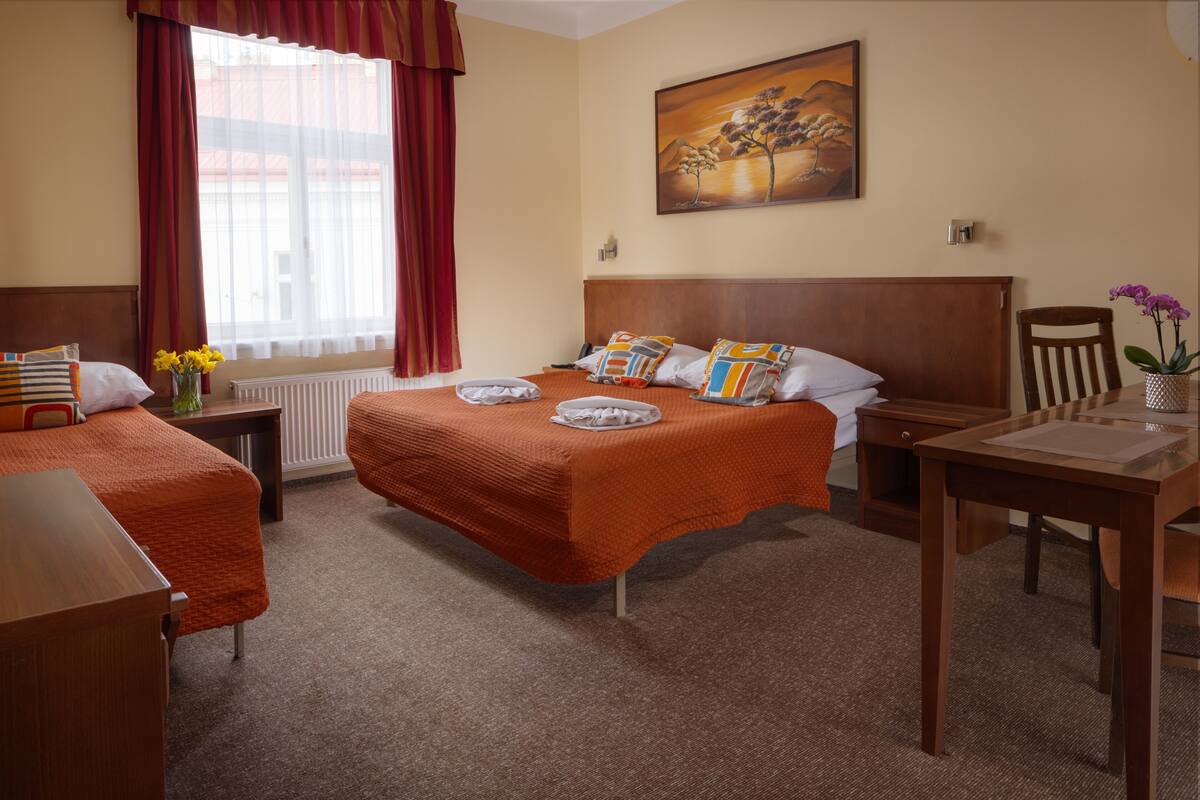 A hotel room with two beds, a dining table, a chair, a radiator, and a painting on the wall.