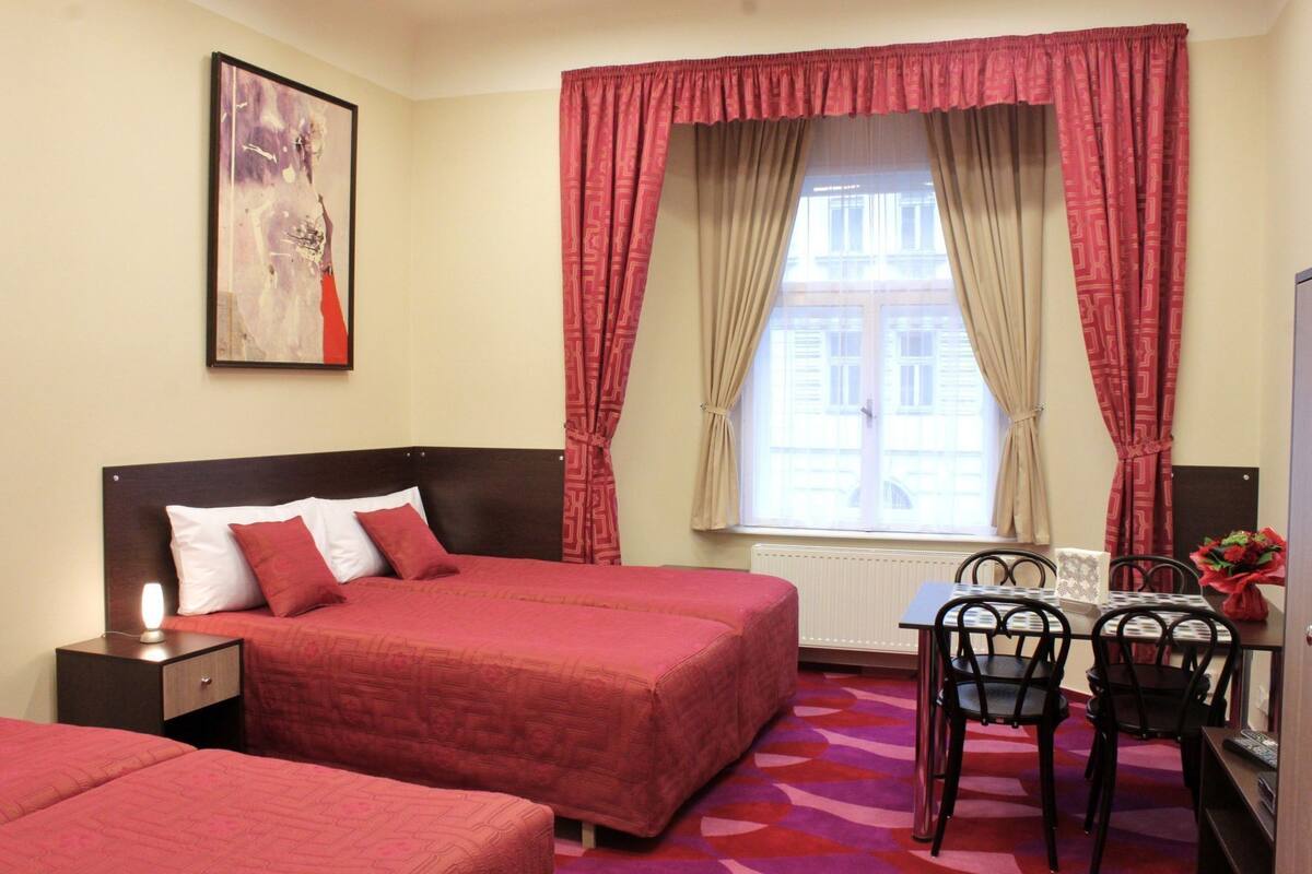 A hotel room with two beds, a desk with chairs, a radiator, and a window with curtains.