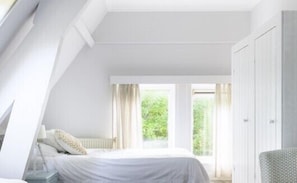 Un dormitorio con techo inclinado, una cama con ropa de cama blanca, una mesita de noche, una ventana con cortinas y un armario.
