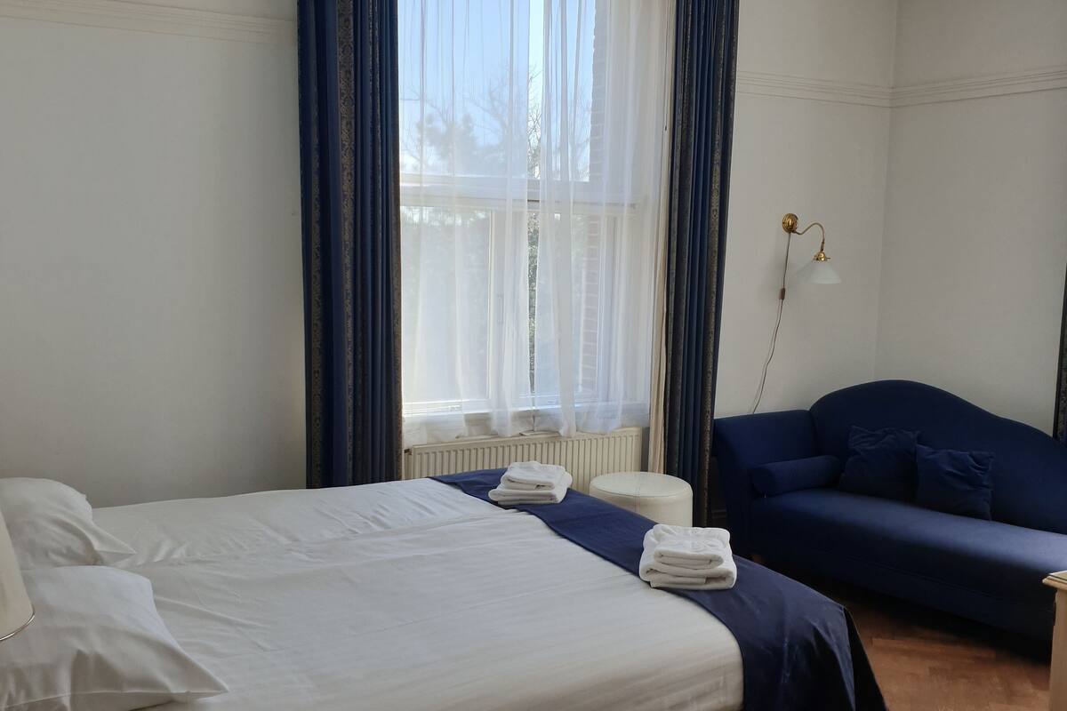 A hotel room with a bed, a sofa, a bedside table, and a window with curtains.