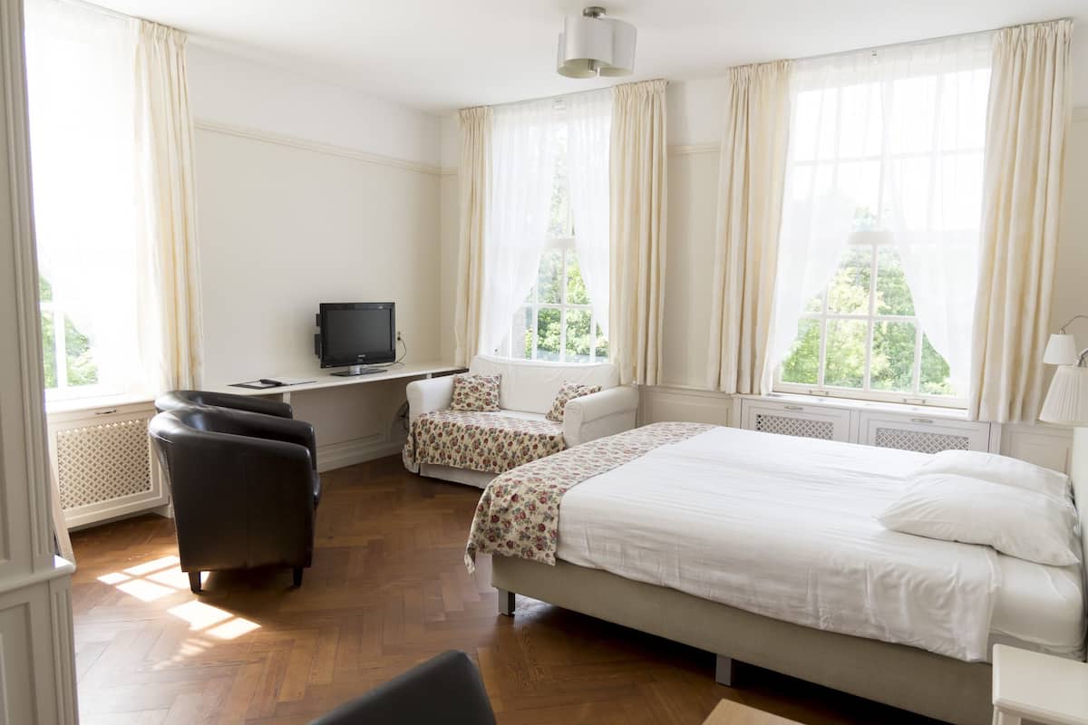 A hotel room with a bed, two chairs, a desk with a television, and large windows with curtains.
