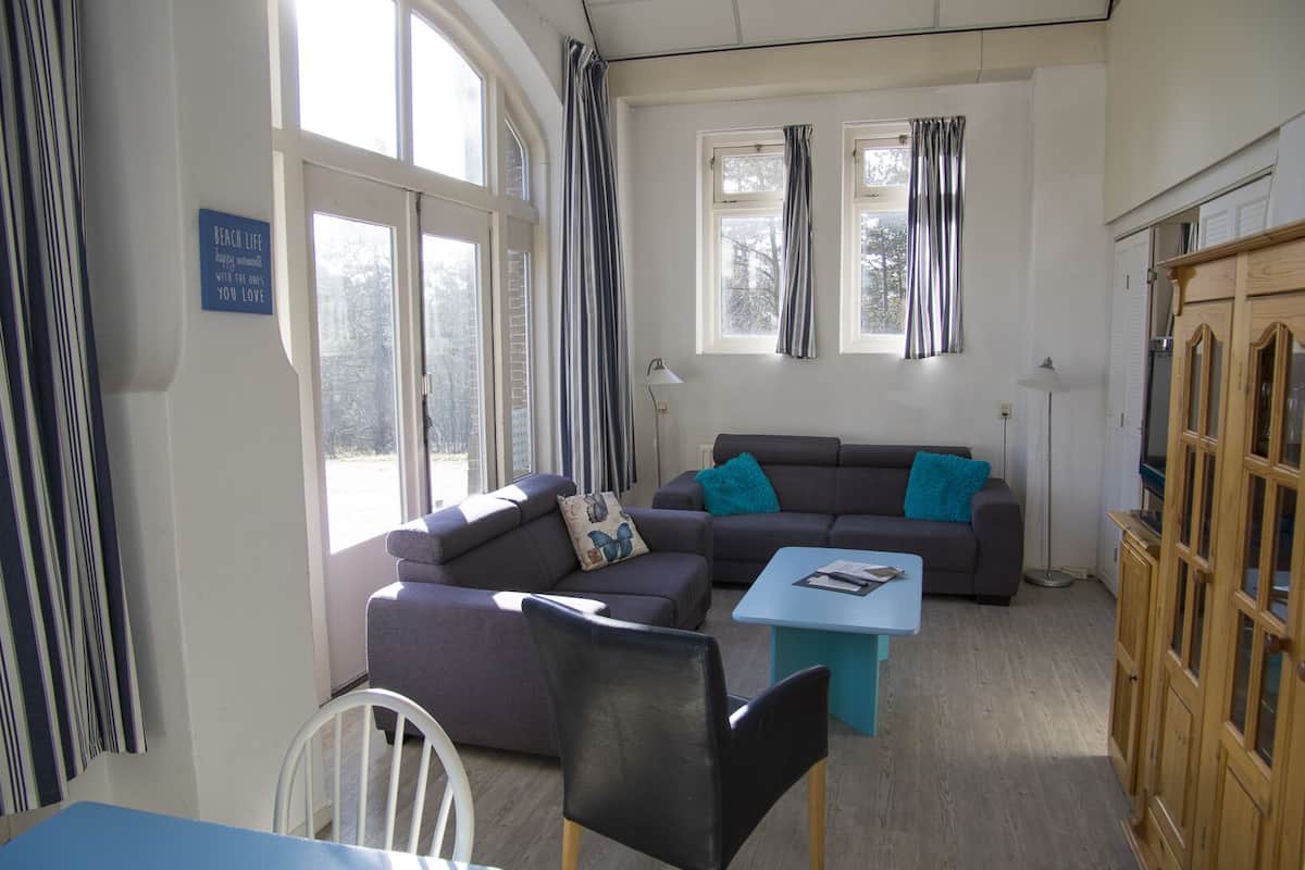 A cozy living room with a sofa set, a coffee table, and a dining table. There are two windows with curtains, a lamp, and a sign on the wall.