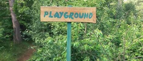 Un letrero de madera con la palabra 'PLAYGROUND', sobre un fondo de follaje verde denso y árboles.