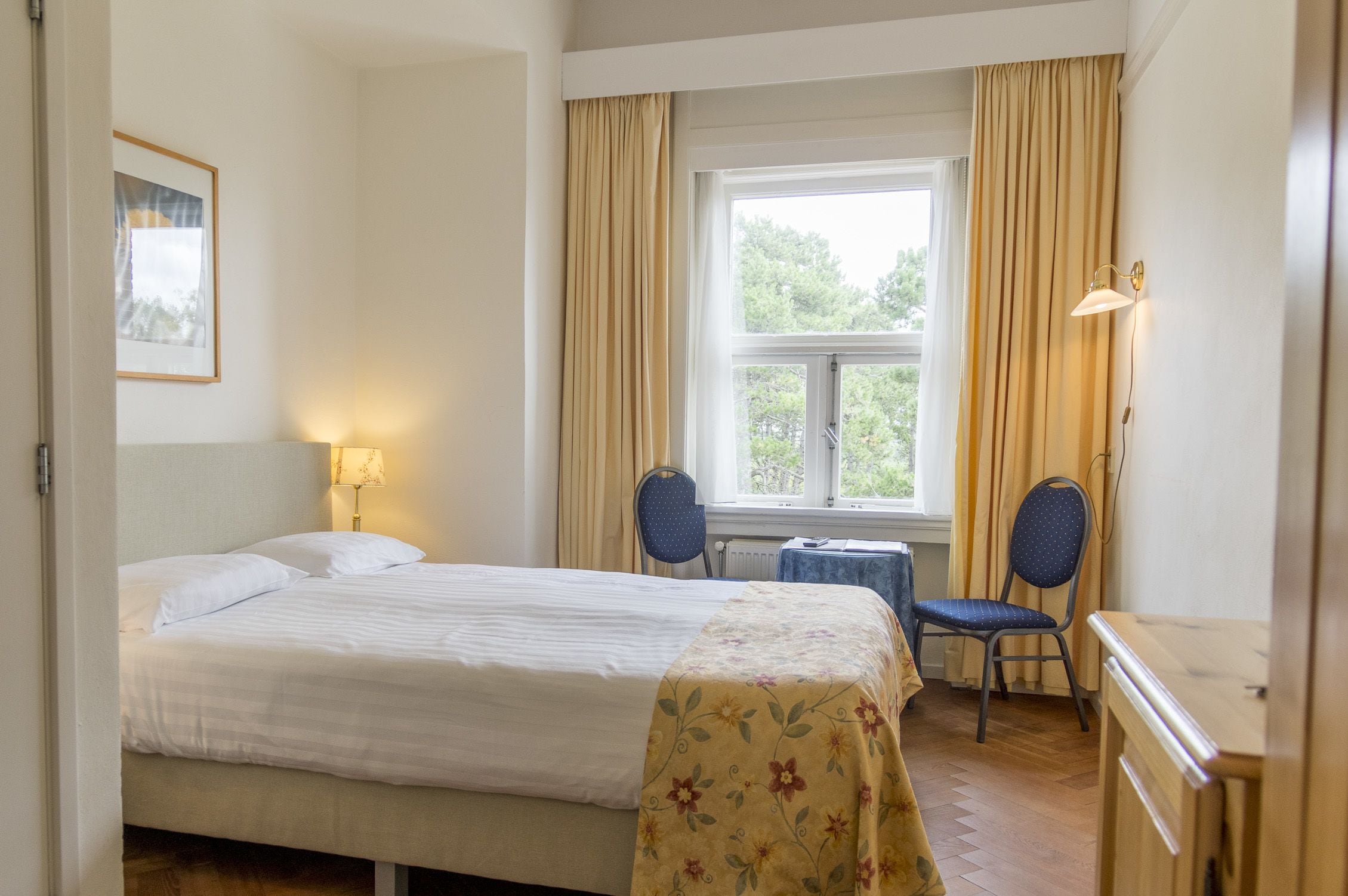 A hotel room with a bed, a desk, two chairs, a window with curtains, a lamp, and a framed picture on the wall.