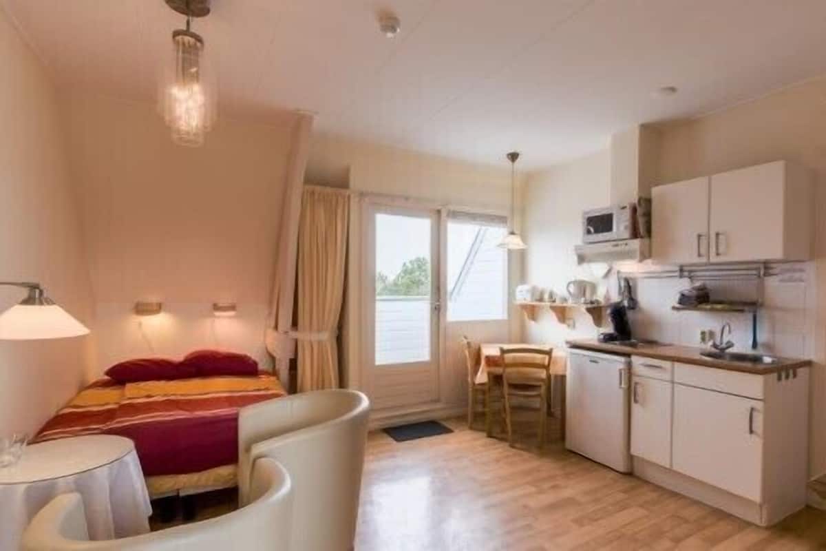 A compact room with a bed, a small table, a chair, a kitchenette, and a window with curtains.