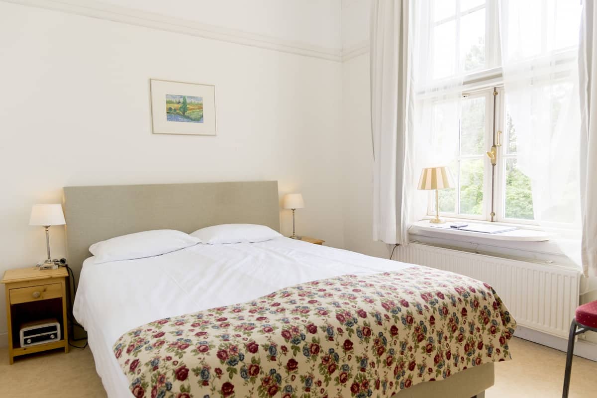A neatly made bed with a floral bedspread, a bedside table with a lamp, and a window with sheer curtains.