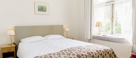 A neatly made bed with a floral bedspread, a bedside table with a lamp, and a window with sheer curtains.