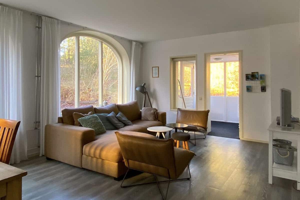 A modern living room with a sectional sofa, a coffee table, and a chair. There are two doors leading to other rooms, and a large window with curtains.