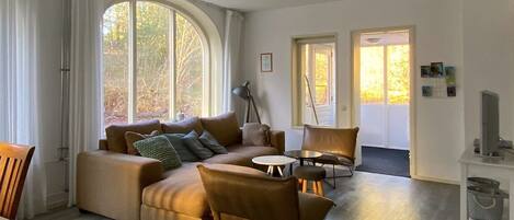 Un salon moderne avec un canapé d’angle, une table basse et un fauteuil. Il y a deux portes qui donnent sur d’autres pièces, ainsi qu’une grande fenêtre avec des rideaux.