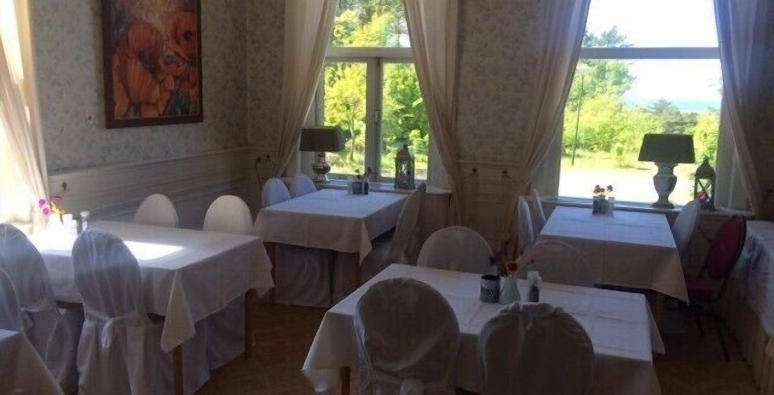 A dining area with tables set for guests, large windows with curtains, and a painting on the wall.
