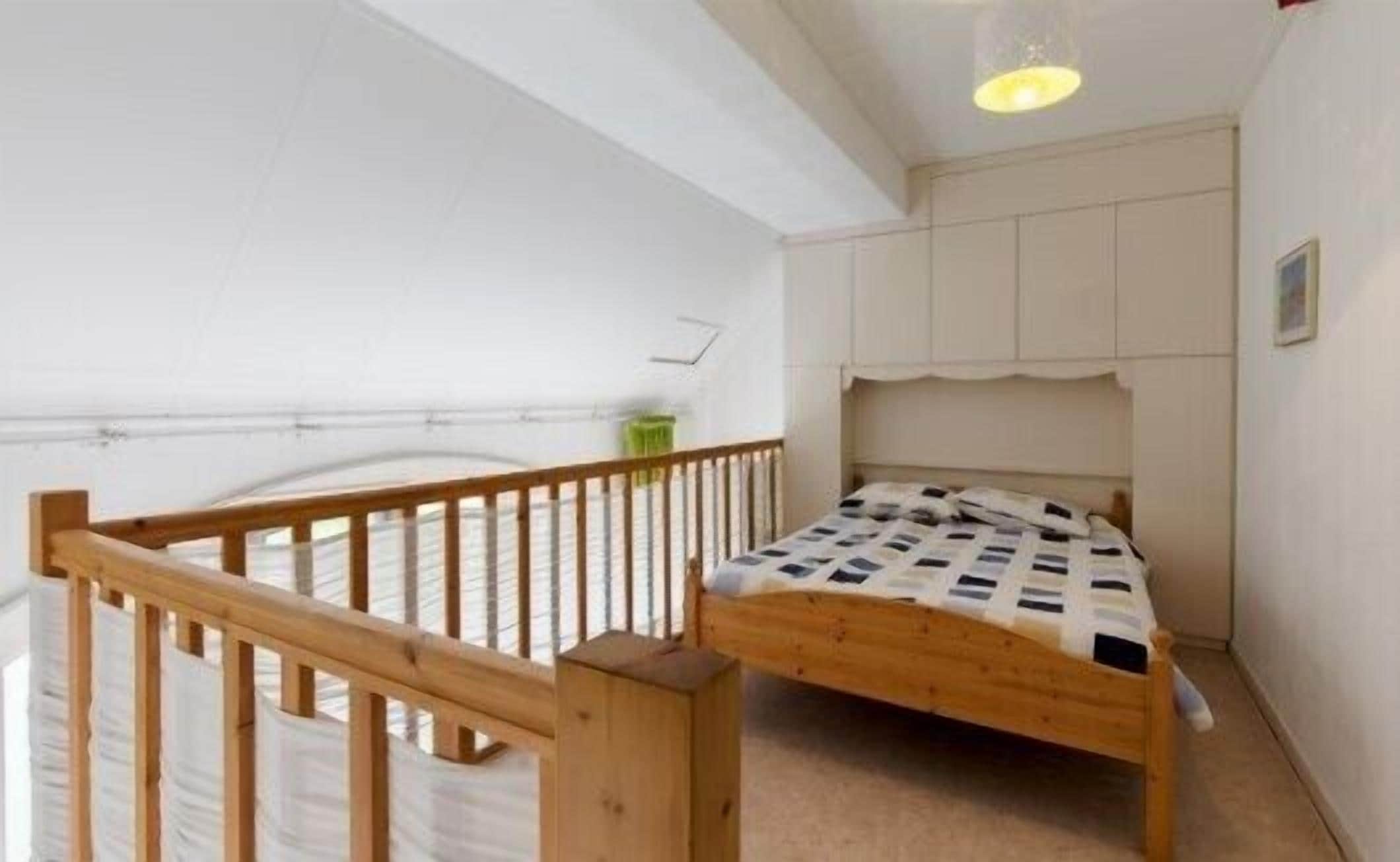 A small, single-bed room with a wooden bed frame, a simple white wall, and a small window.