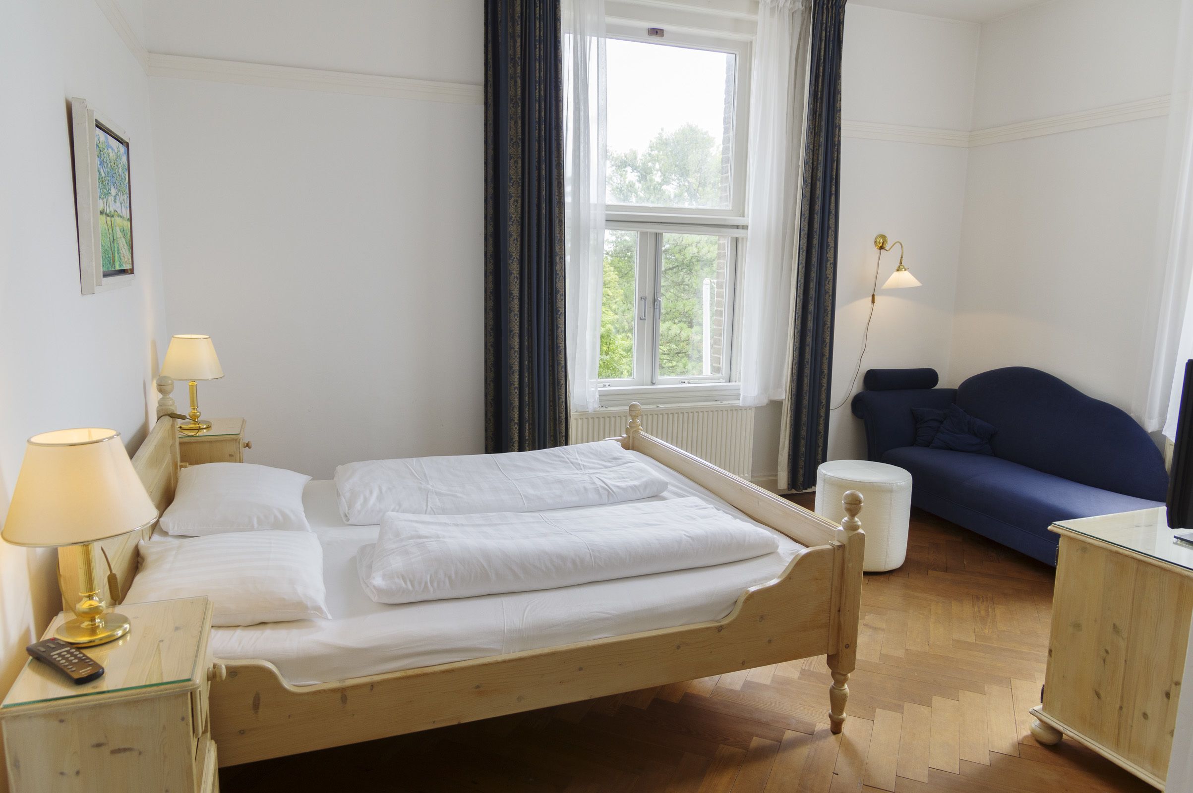 En snyggt bäddad säng med vita sängkläder, en sänglampa, en fjärrkontroll och ett litet sidobord. I rummet finns ett fönster med gardiner, en blå soffa och ett träskåp.