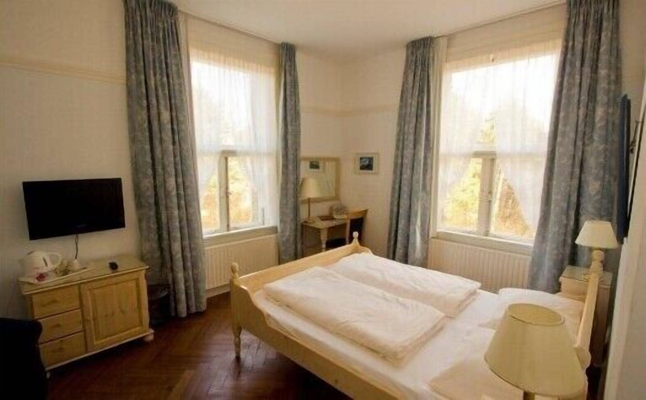 A bedroom with a wooden bed, a bedside table, a lamp, a TV, and a window with curtains.