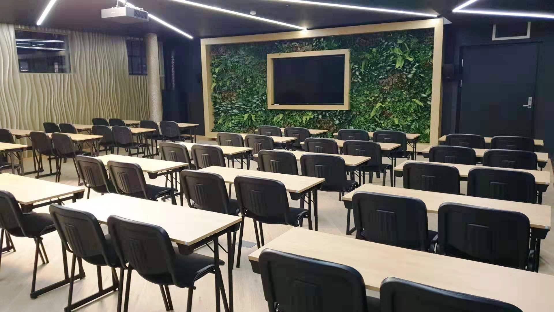 Un aula con filas de escritorios y sillas, una pantalla grande y una pared verde con follaje.