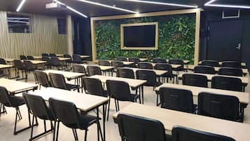 Un aula con filas de escritorios y sillas, una pantalla grande y una pared verde con follaje.
