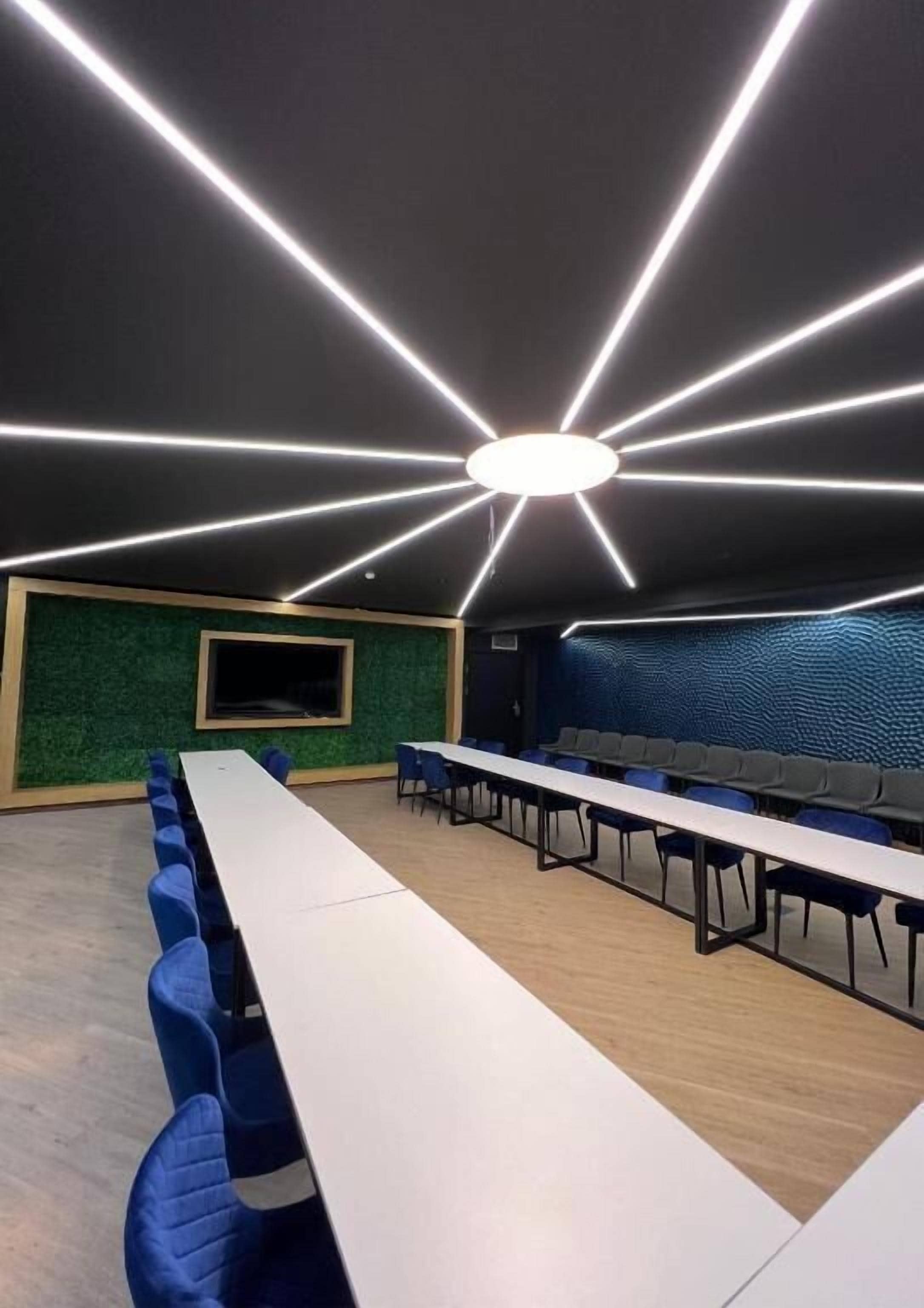 A conference room with a long table, blue chairs, a large screen, and a modern ceiling light design.