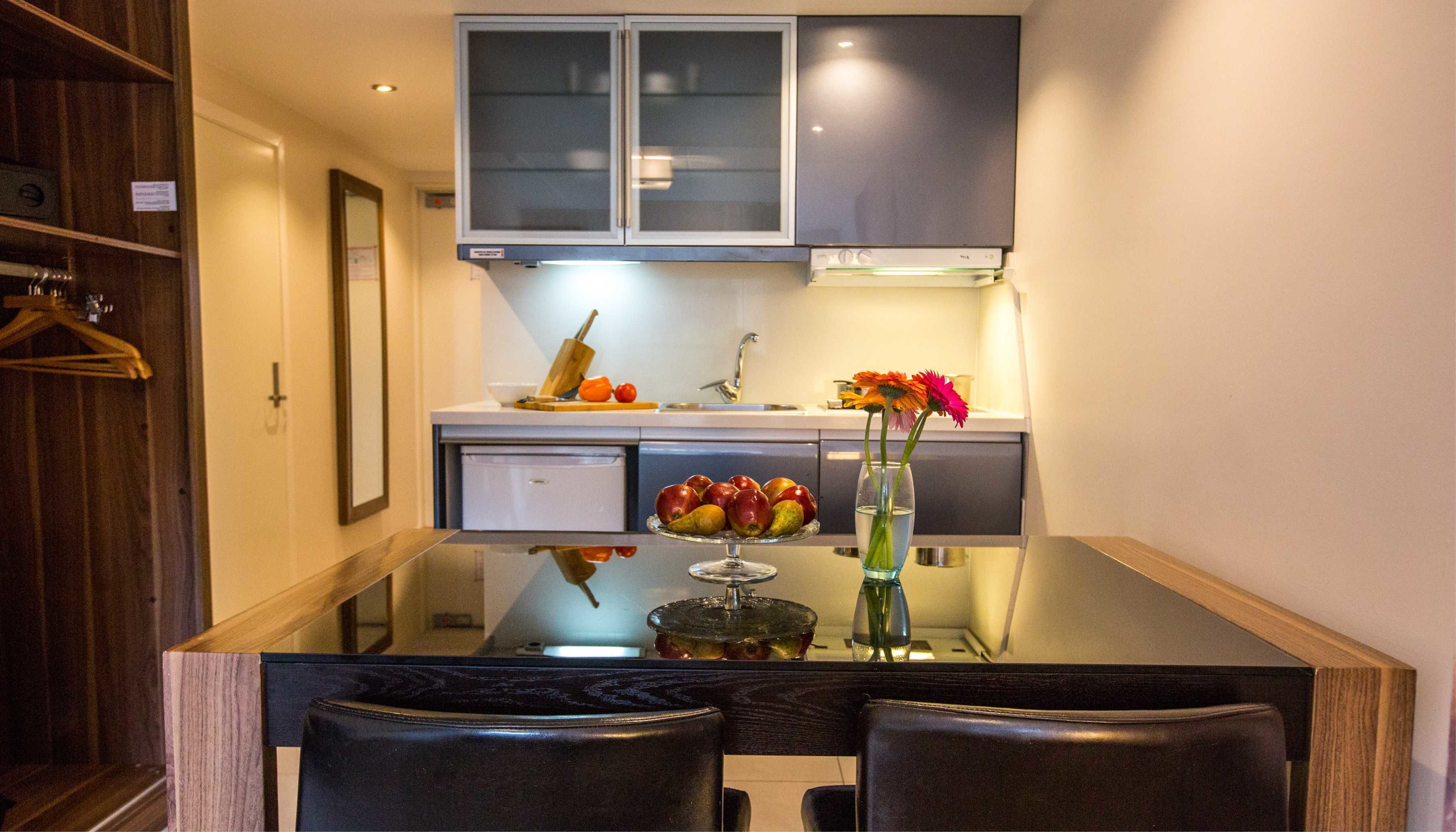 Een moderne keuken met een glazen eettafel, donkere kastjes en een kleine koelkast.