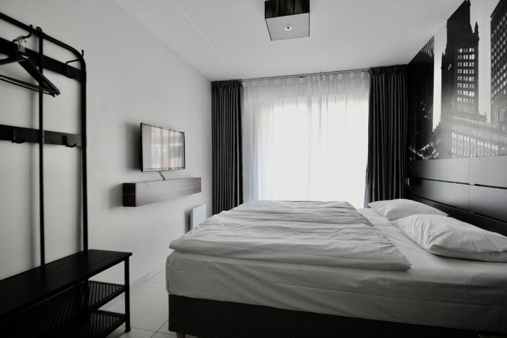 A modern hotel room with a bed, a TV mounted on the wall, a bench, and a window with curtains.