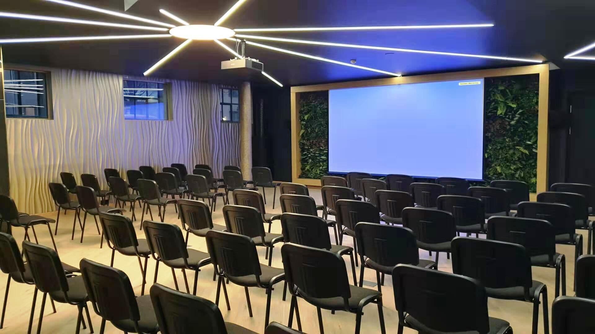 A conference room with rows of black chairs, a large screen, and a modern ceiling light fixture.