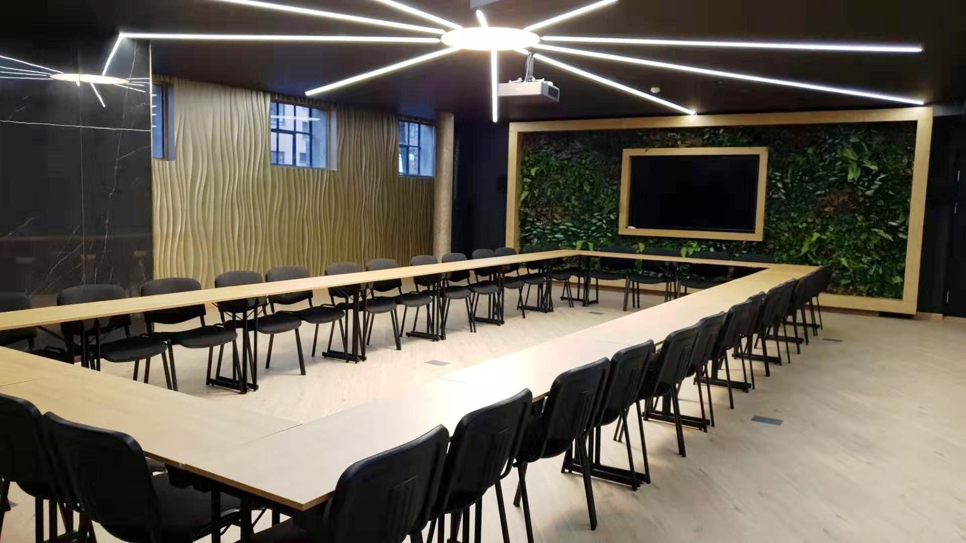 A conference room with a long table, black chairs, a projector screen, and a green wall with plants.