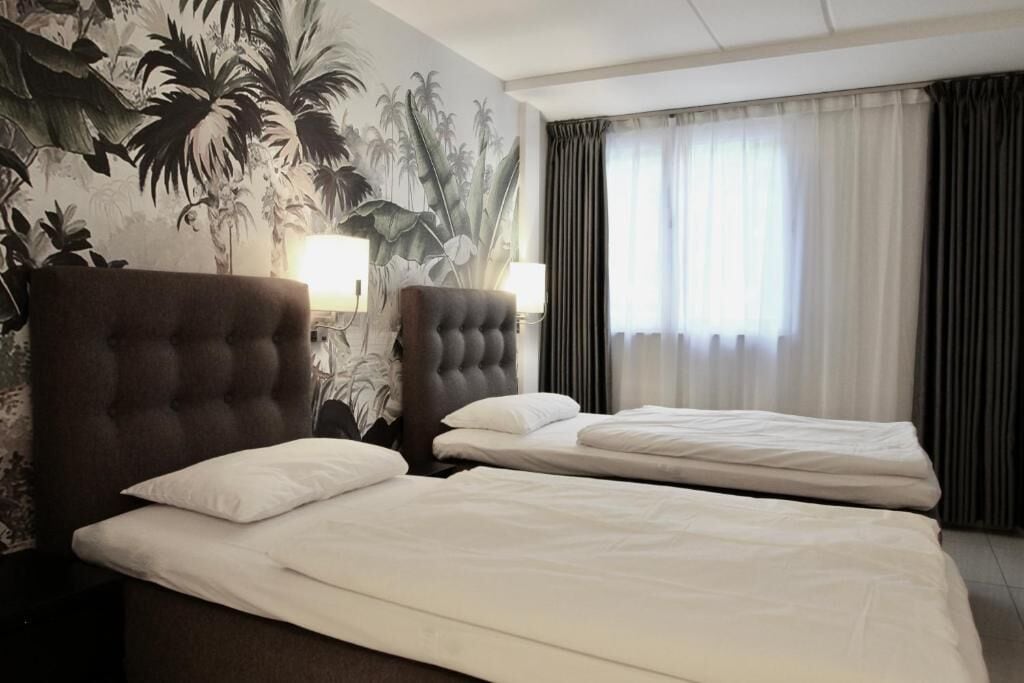 Ett hotellrum med två sängar, en stor väggmålning med tropiska växter och utsikt över omgivningen genom ett fönster med gardiner.