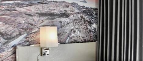 Una habitación de hotel con cama, mesita de noche, lámpara, un mural grande y una cortina.