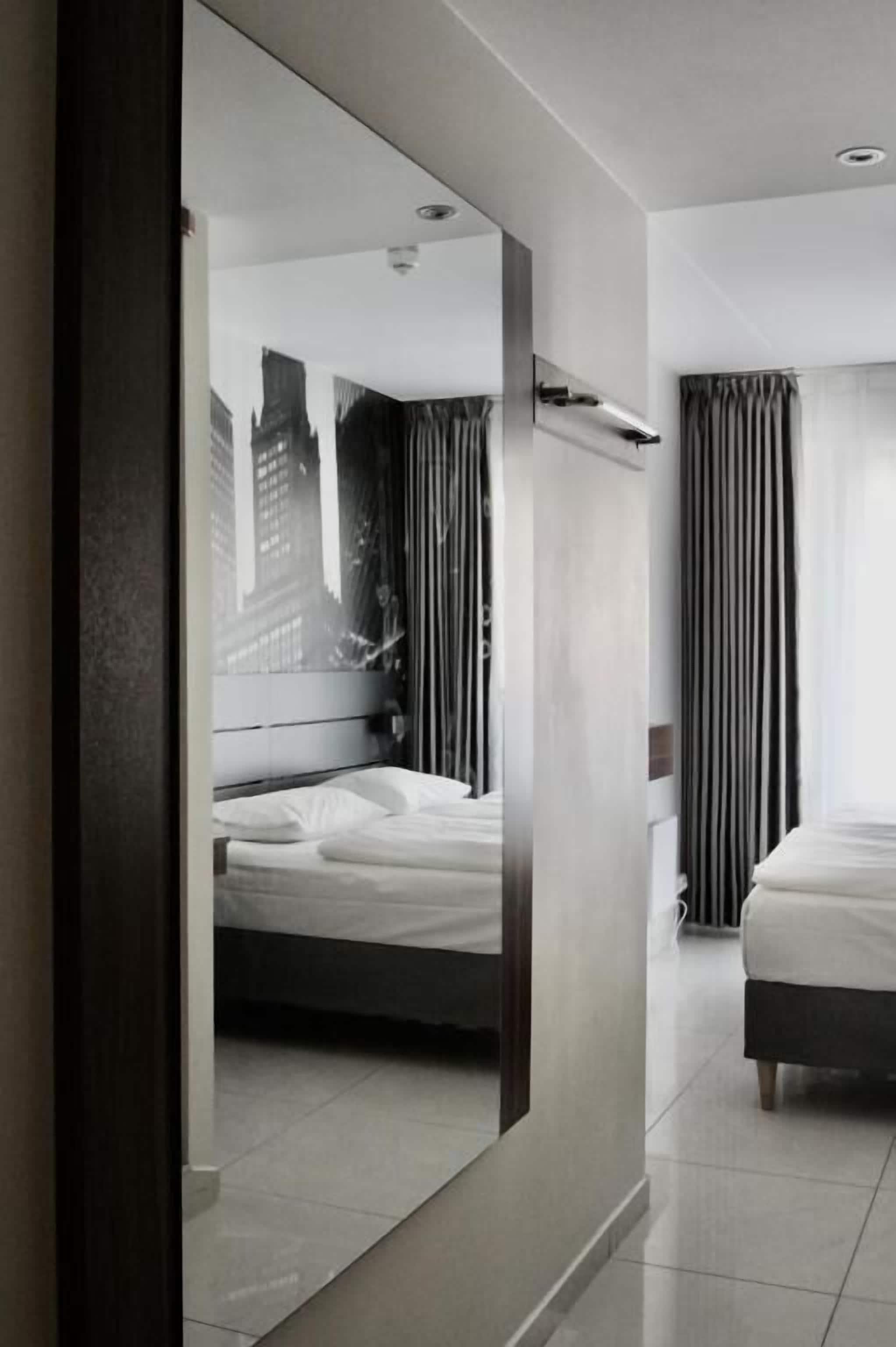 A modern hotel room with a large mirror reflecting a cityscape, a bed with white linens, a bedside table, and a window with curtains.