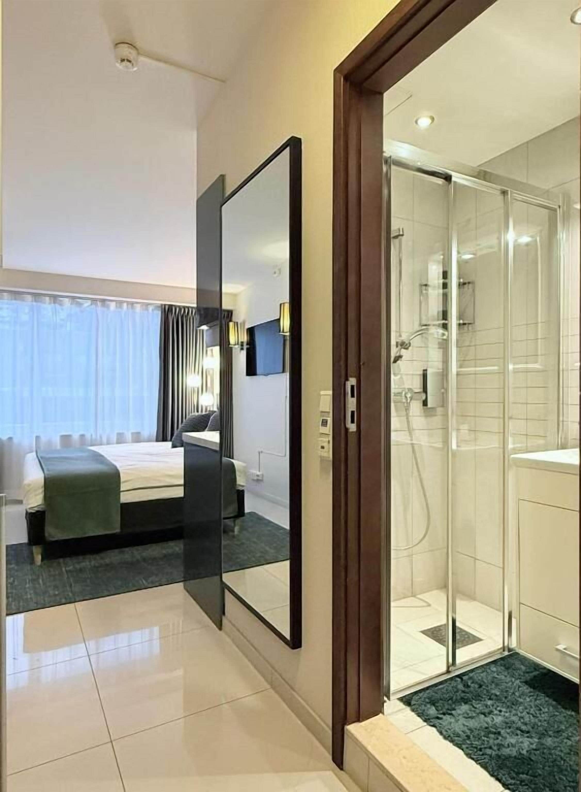 A hotel room with a bed, a large mirror, a shower area, and a view of the room through the mirror.