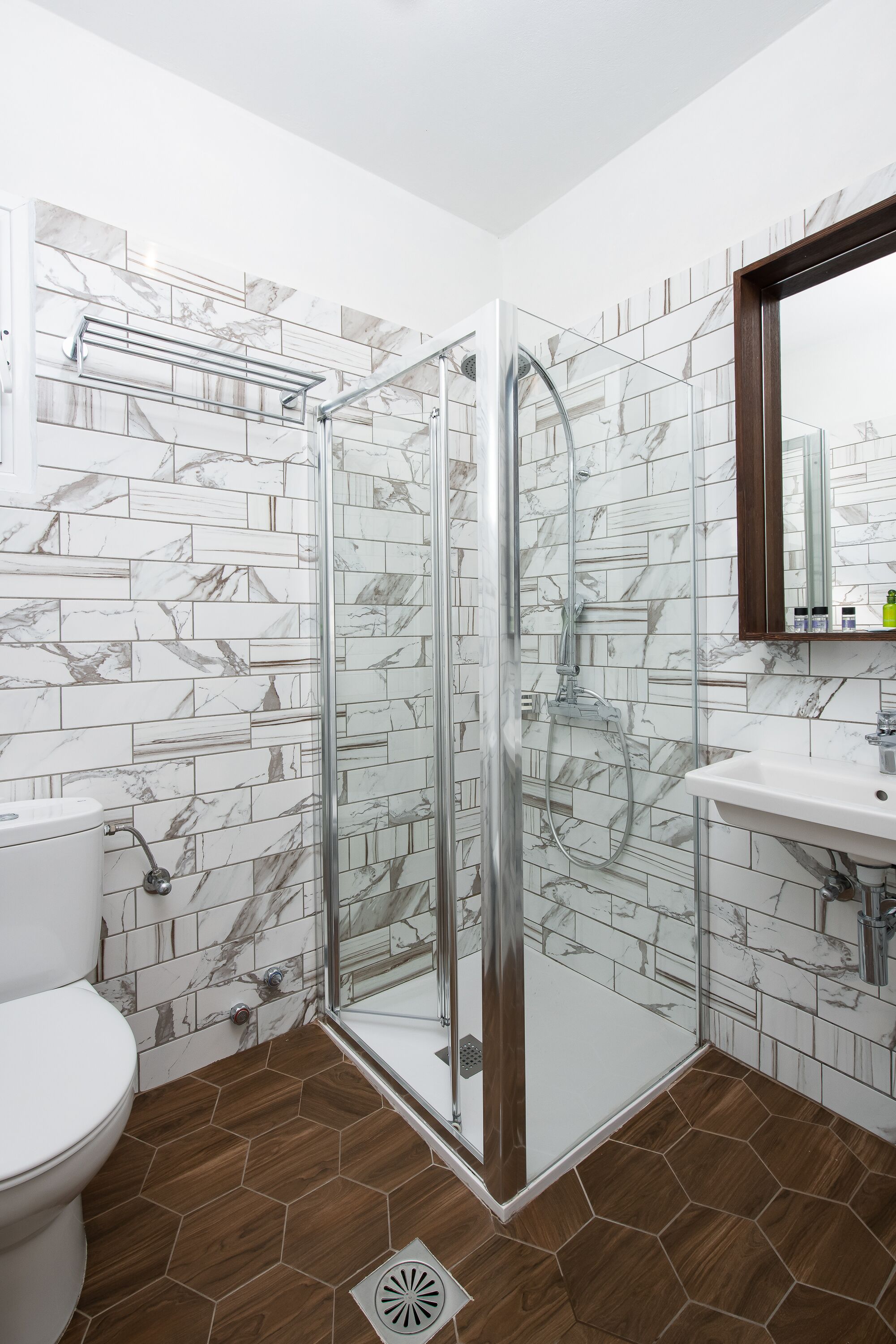 Une salle de bain dotée d’une douche à parois de verre, de toilettes blanches, d’un lavabo avec un miroir et de carreaux aux motifs de marbre sur les murs.