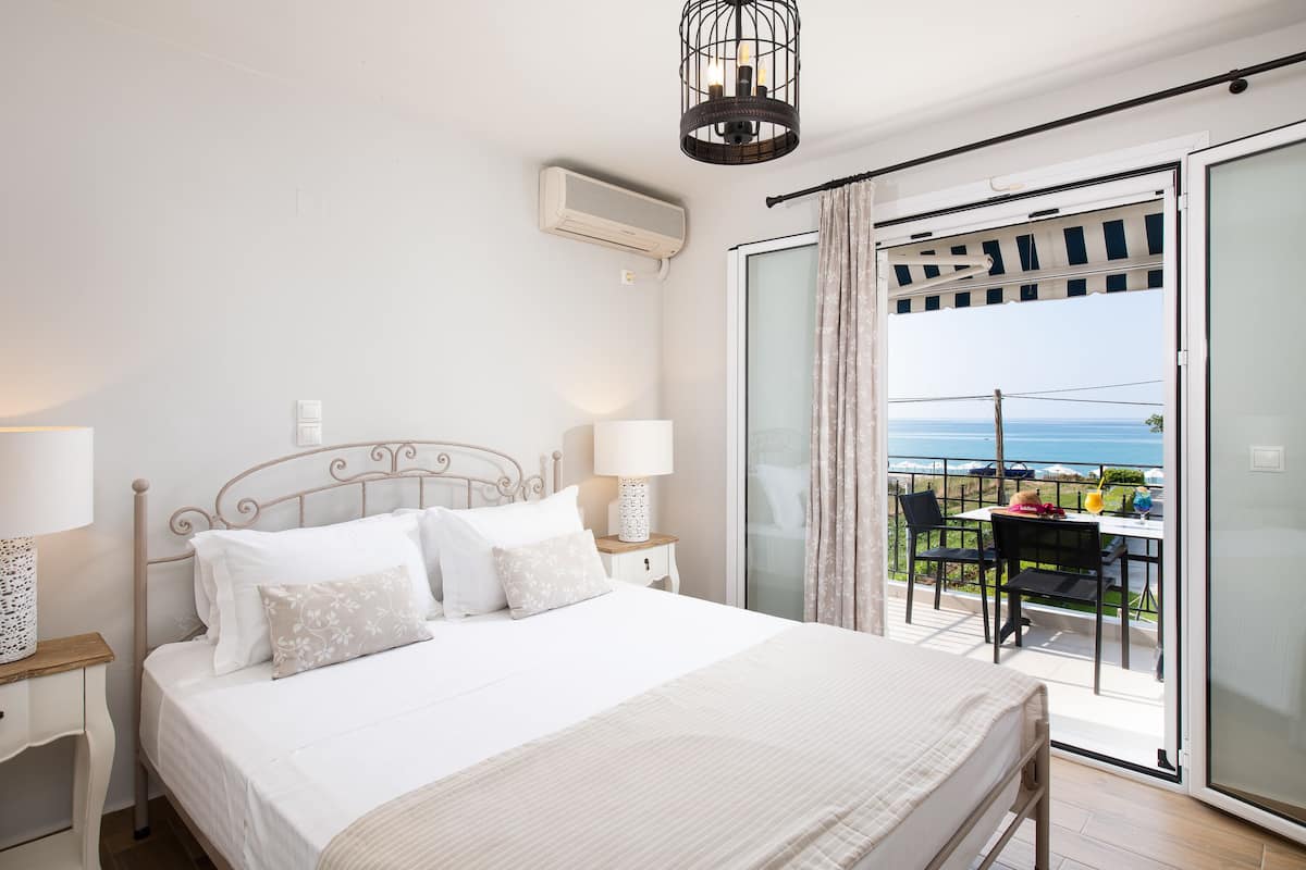 A bedroom with a bed, bedside lamps, an air conditioner, and a view of the sea through a sliding glass door.