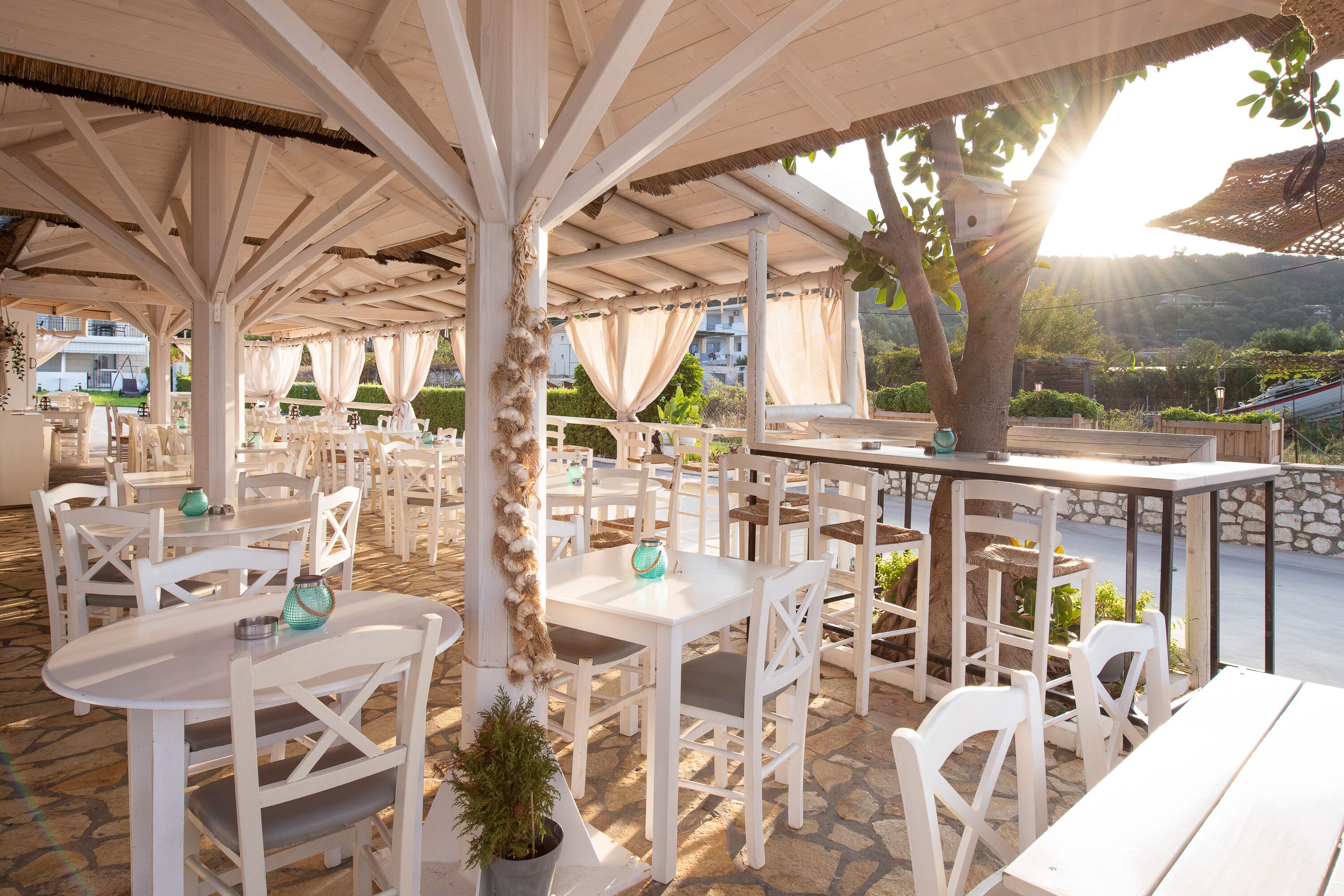 An outdoor patio with white tables and chairs, a thatched roof, and a view of the surrounding landscape.
