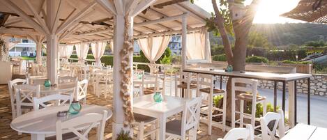 An outdoor patio with white tables and chairs, a thatched roof, and a view of the surrounding landscape.