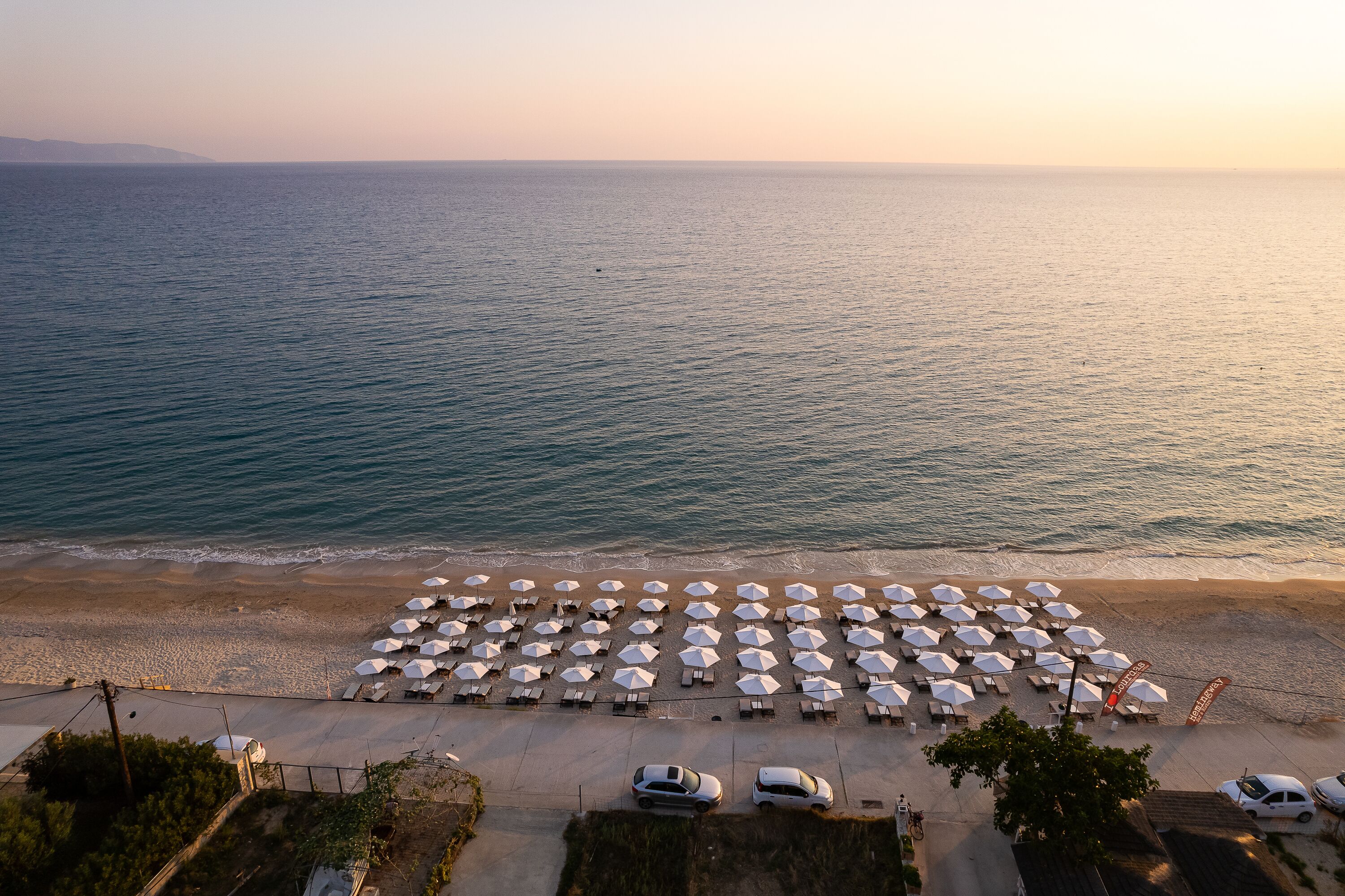 Une vue aérienne d’une plage avec des rangées de parasols et de chaises de plage, un parking avec des voitures, et une vue dégagée sur la mer.