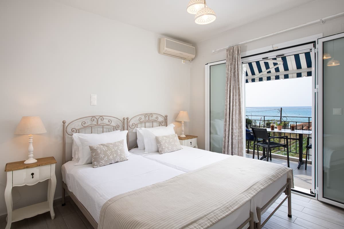 A bedroom with a large bed, two bedside tables with lamps, a ceiling fan, and a sliding glass door leading to a balcony with a view of the sea.