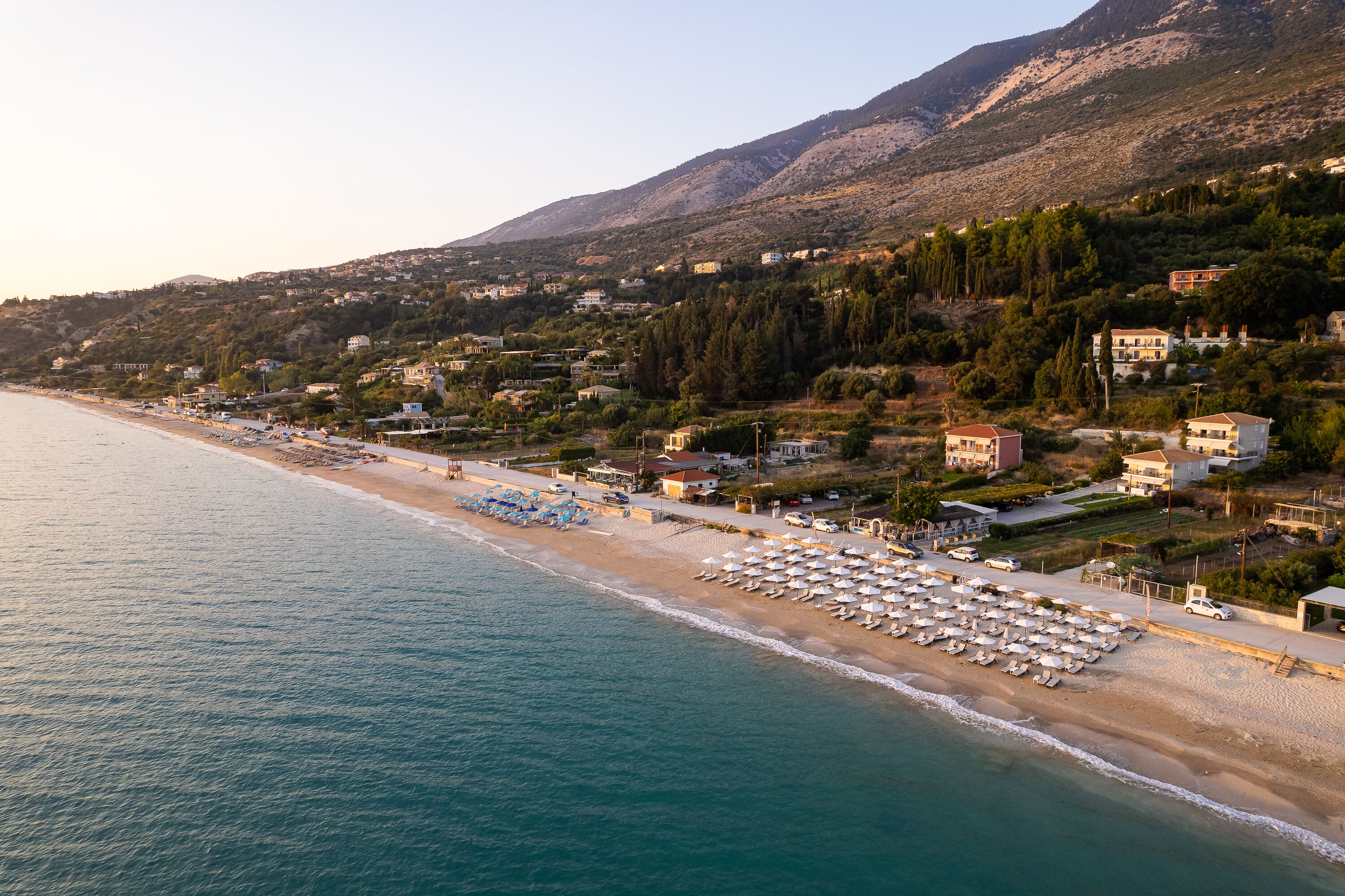 A coastal town with a beach lined with umbrellas and sun loungers, houses on a hillside, and a mountain in the background.