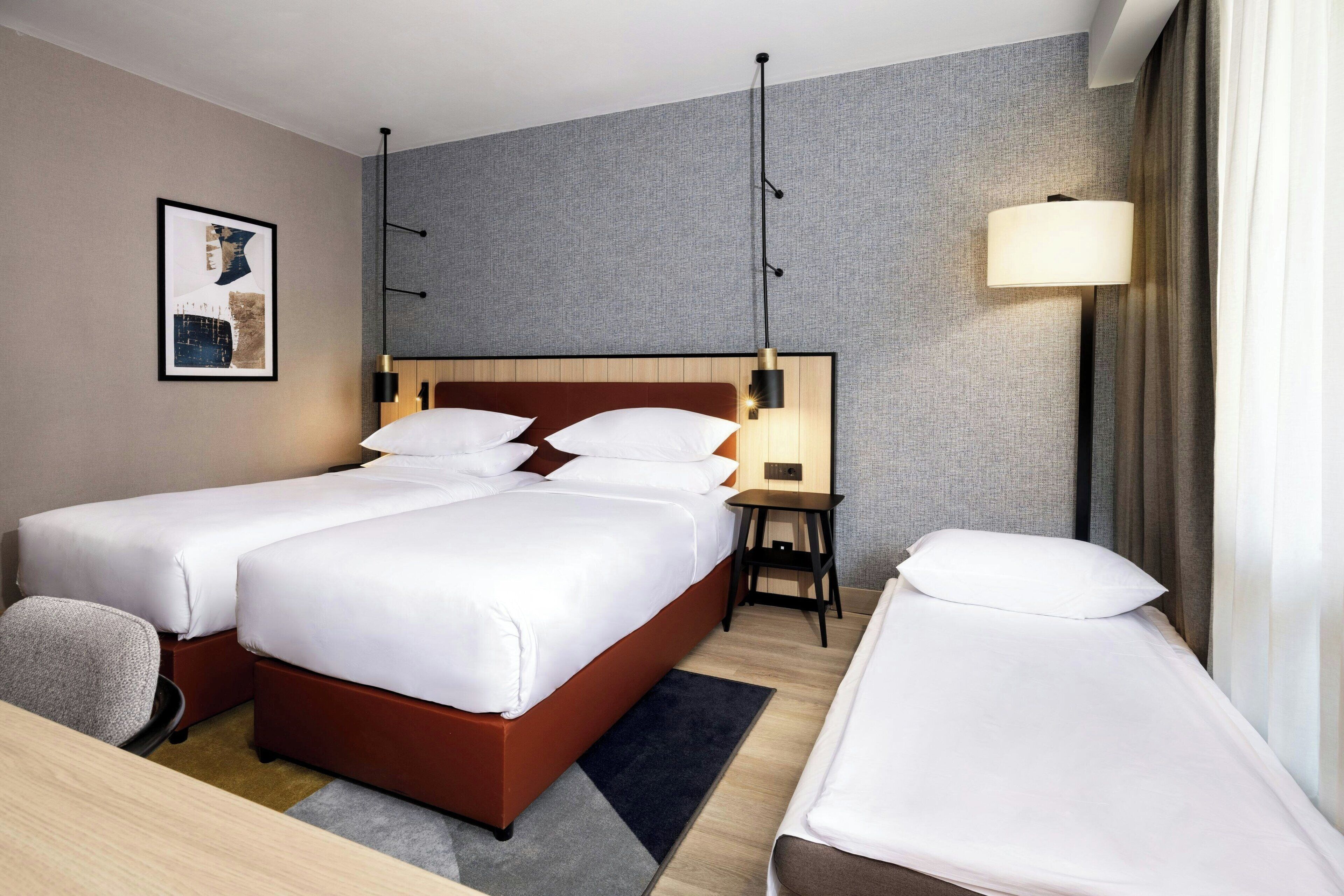 A hotel room with two beds, a desk, a chair, a lamp, and a framed picture on the wall.