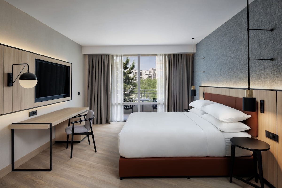 Une chambre d’hôtel moderne dotée d’un grand lit, d’un bureau, d’une chaise, d’un téléviseur fixé au mur et d’un balcon offrant une vue sur la ville.