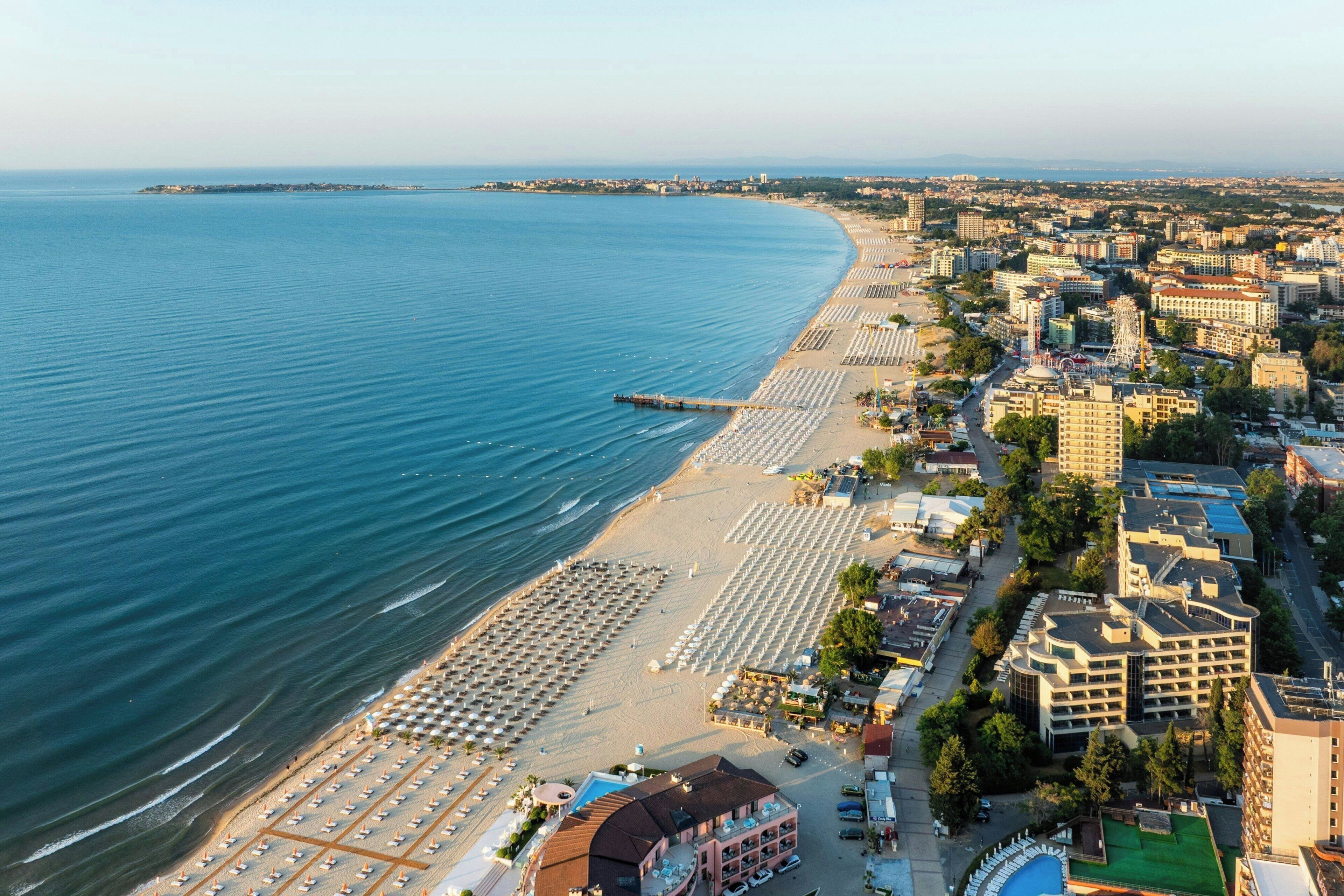 A coastal cityscape with a beach lined with rows of sunbeds and umbrellas, a pier extending into the sea, and a mix of residential and commercial buildings.