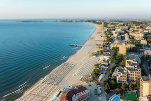 Un paisaje urbano costero con una playa bordeada de filas de tumbonas y sombrillas, un muelle que se adentra en el mar, y una mezcla de edificios residenciales y comerciales.