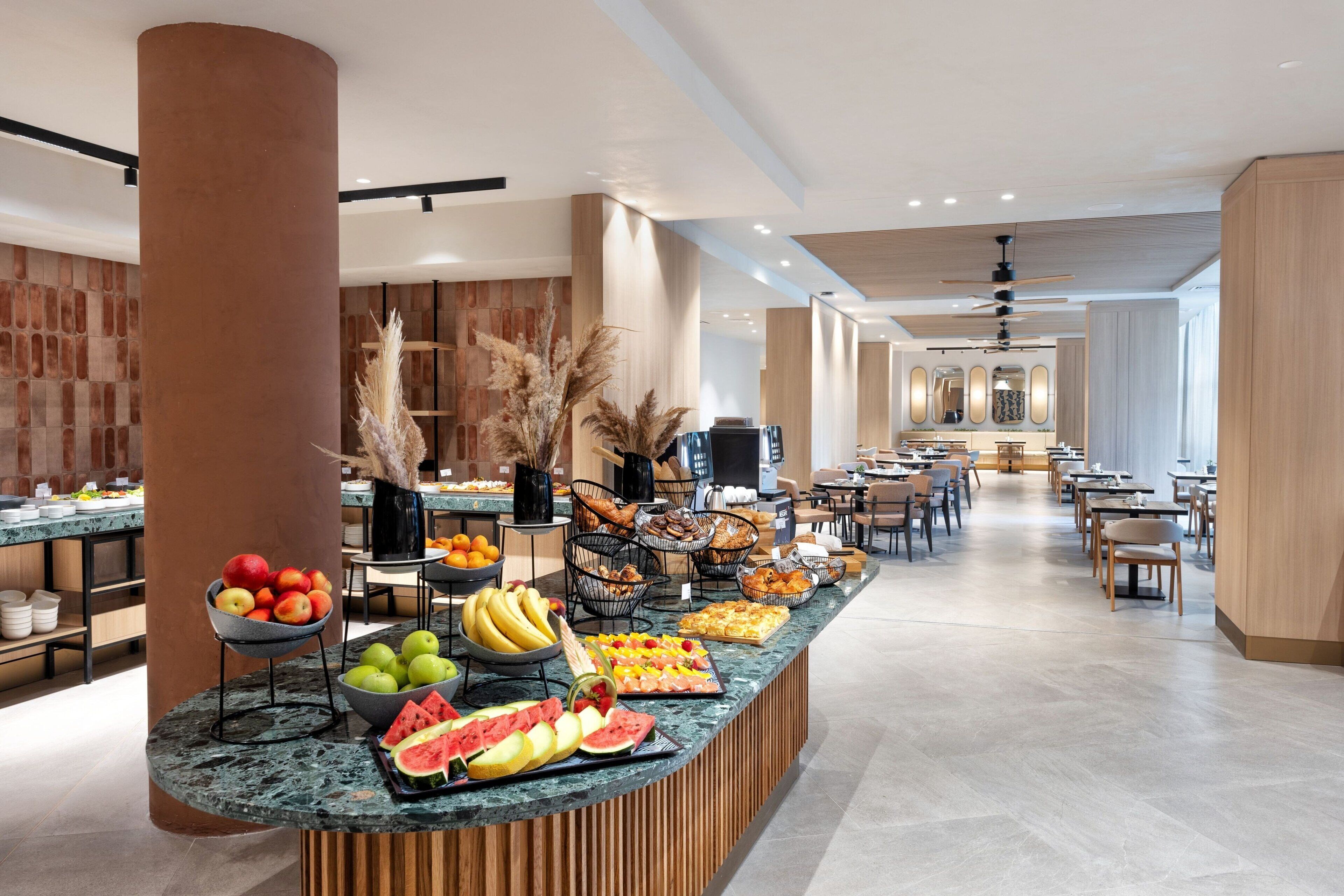 A modern hotel lobby with a central counter displaying fresh fruits and pastries, wooden accents, and a spacious seating area.
