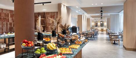 A modern hotel lobby with a central counter displaying fresh fruits and pastries, wooden accents, and a spacious seating area.