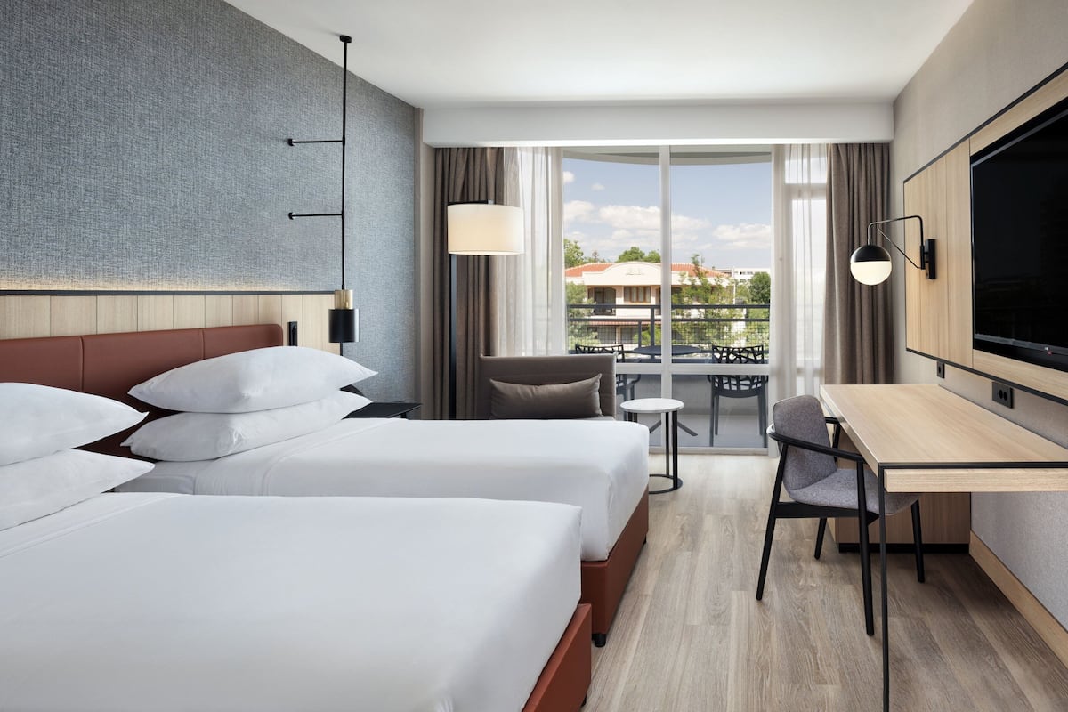 Une chambre d’hôtel avec deux lits, un bureau, une chaise, une télévision et un balcon donnant sur des immeubles.