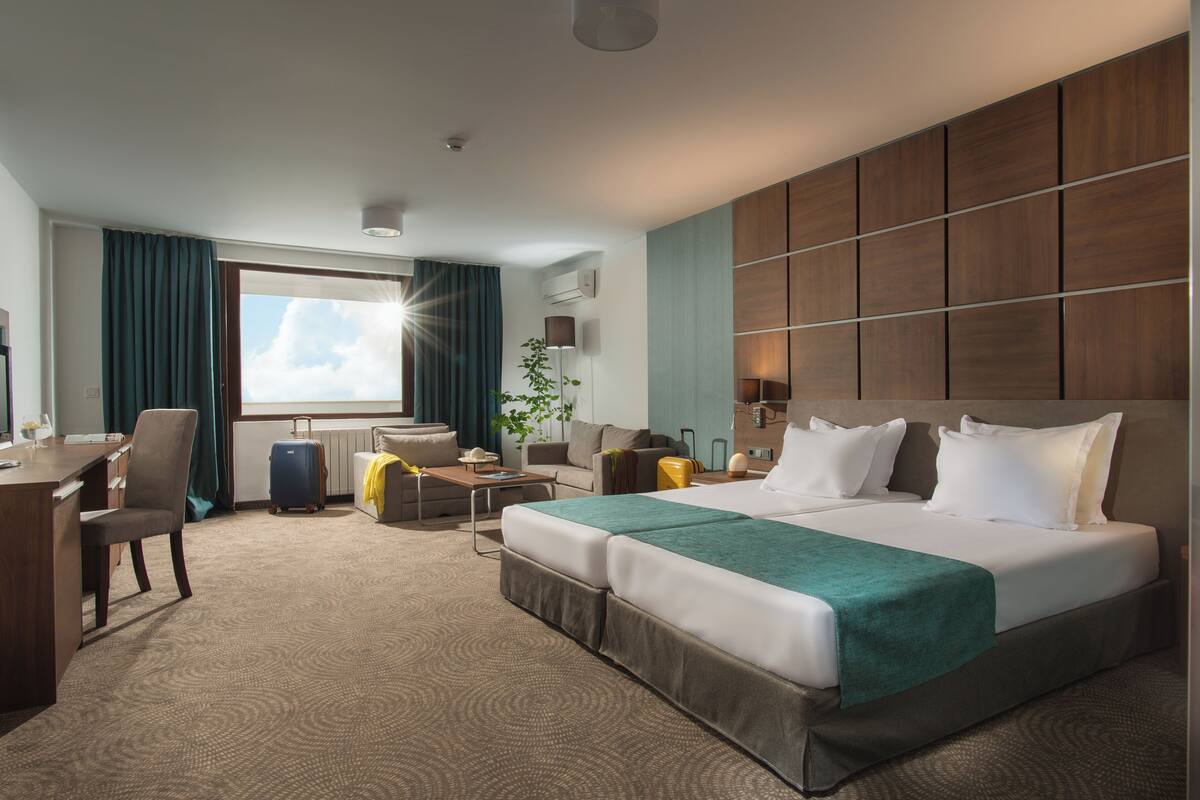 A hotel room with a large bed, a desk, a chair, a small table, a plant, and a window with curtains.