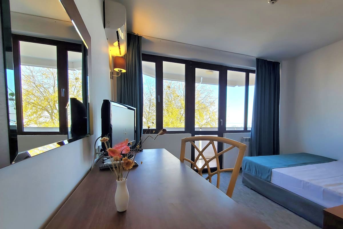 A hotel room with a bed, a desk with a computer, a chair, a TV, and large windows with curtains.