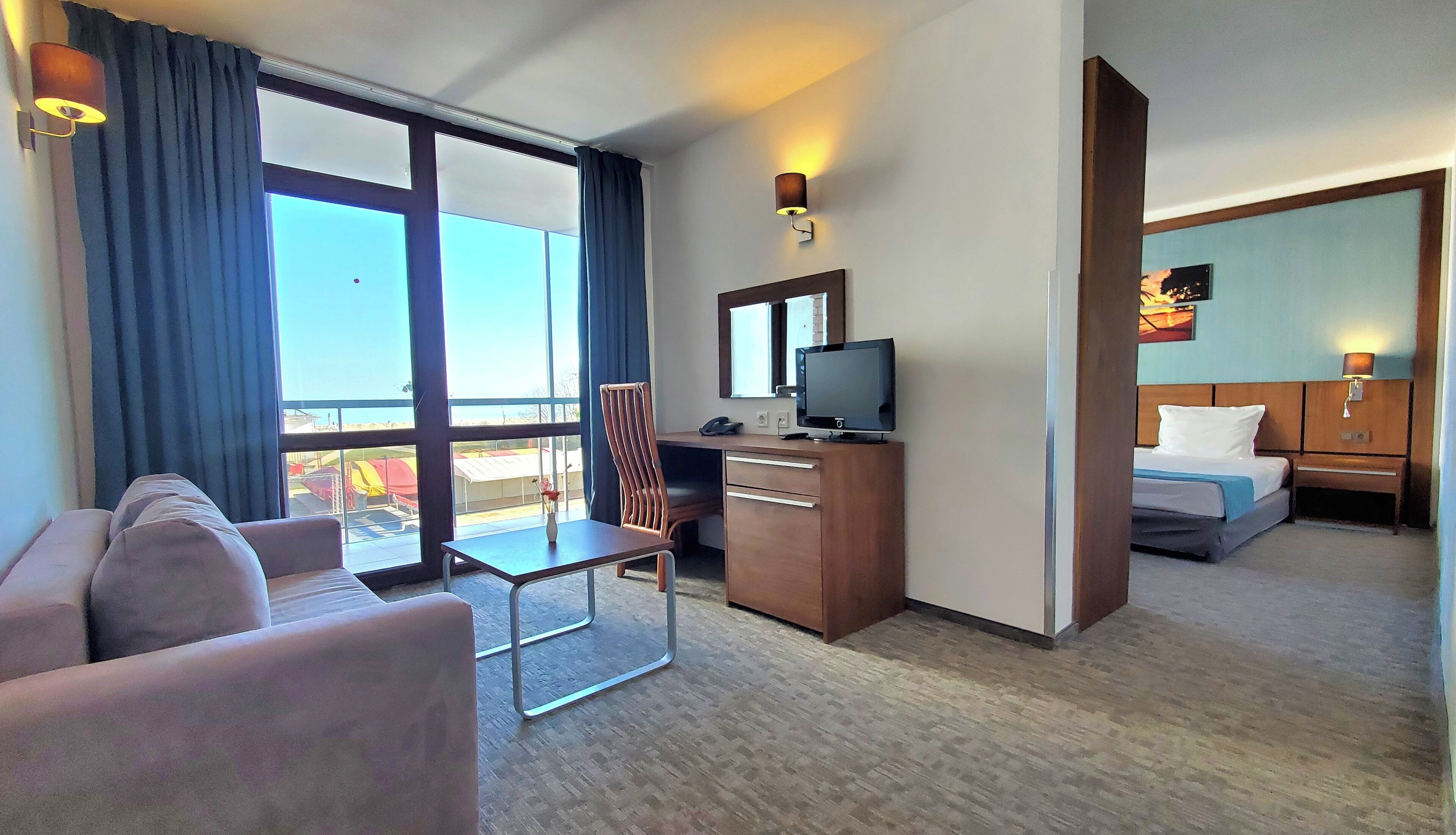 A hotel room with a sofa, a desk with a TV, a chair, a bed, and a large window with curtains.